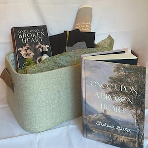 May include: A gift basket filled with books, stationery, and a small bottle. The books are titled "Once Upon a Broken Heart" by Stephanie Garber. The basket is light green with brown leather handles. The items are arranged on a white surface.