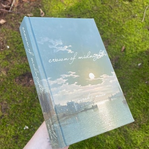 Puede incluir: Un libro de tapa dura titulado "Crown of Midnight" de Sarah J. Maas. La portada presenta un paisaje sereno con una luna, nubes y agua. El libro se sostiene sobre un fondo de follaje verde.