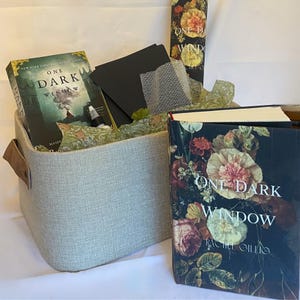 Op de afbeelding: Een cadeaupakket met het boek "One Dark Window" van Rachel Gillig, met een bloemenomslag. De mand bevat een bijpassende boekenrol, zwarte notitiekaarten en een pen. De lichtgrijze mand heeft bruine leren handvatten. Het boek maakt deel uit van de New York Times Bestseller-serie.
