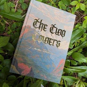 Peut inclure: Un livre relié intitulé The Two Towers de J.R.R. Tolkien. La couverture présente une illustration de paysage avec des montagnes bleues et orange sous un ciel nuageux. Le titre est imprimé dans une grande police gothique sombre.