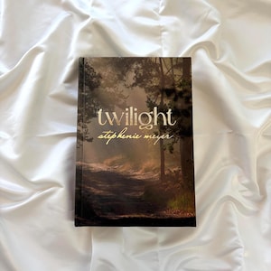 Puede incluir: Un libro de tapa dura titulado Twilight de Stephenie Meyer con una escena de bosque oscuro en la portada. La ilustración muestra árboles altos, un camino de tierra y luz dorada filtrándose entre las ramas. El título y el autor están en oro.