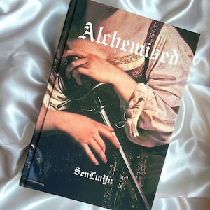 May include: A hardcover book titled "Alchemized" with a Renaissance-style painting on the cover. The artwork depicts a person holding a sword. The author's name, "SenLinyu," is printed at the bottom in a gothic font. The book rests on a white satin surface.