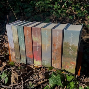 Pode incluir: Uma coleção de livros de capa dura com capas coloridas, com títulos como "Kingdom of Ash" e "Tower of Dawn". As lombadas exibem uma escrita elegante e ilustrações de paisagens, sugerindo um gênero de fantasia ou ficção histórica. Os livros estão dispostos ao ar livre.