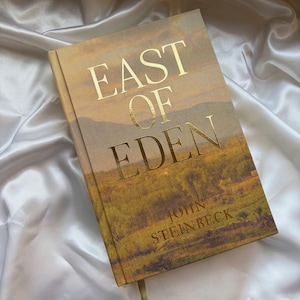 Może przedstawiać: Książka w twardej oprawie zatytułowana "East of Eden" autorstwa Johna Steinbecka. Okładka książki zawiera złote litery na tle ilustracji krajobrazu w odcieniach brązu, zieleni i żółci. Książka spoczywa na białej, jedwabistej powierzchni.