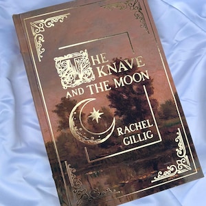 Op de afbeelding: Een hardcover boek getiteld The Knave and the Moon door Rachel Gillig. De kaft toont een donker landschapsschilderij met goudfolie accenten, waaronder een maansikkel, een ster, sierlijke hoekranden en elegante letters in een schreeflettertype.