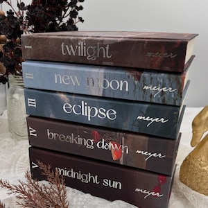 Könnte beinhalten: Ein Stapel aus fünf gebundenen Büchern mit den Titeln Twilight, New Moon, Eclipse, Breaking Dawn und Midnight Sun von Meyer. Jeder Buchrücken zeigt eine römische Ziffer von I bis V, silberne Schrift und dunkle Motive mit roten Früchten und Tropfen.