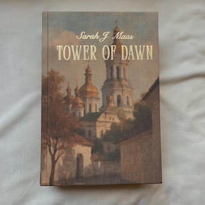 Könnte beinhalten: Ein Hardcover-Buch mit dem Titel "TOWER OF DAWN" von Sarah J. Maas. Das Cover zeigt ein Gemälde einer Stadt mit goldenen Kuppelgebäuden und einem hohen Turm. Die Farbpalette umfasst Beige, Braun und Gold.