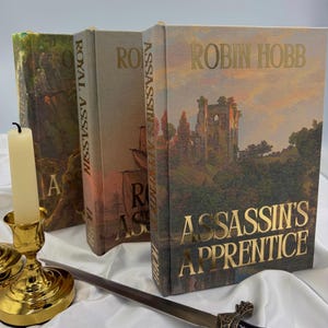 May include: Three hardcover books by Robin Hobb, including Assassin's Apprentice and Royal Assassin, featuring landscape cover art. A gold-colored metal candlestick with a tall cream-colored candle and a metal sword are positioned in the foreground.