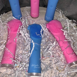May include: Five electric facial cleansing brushes, two blue and three pink, with silicone bristles. The brushes are arranged in a circle on a white surface with blue and silver crinkle paper. The brushes have the text "Nicole Cosmetics" printed on them.
