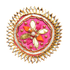 Puede incluir: Un adorno circular de tela rosa con un diseño de flor dorada y naranja. La flor tiene un centro de perla y está rodeada por un borde plateado y dorado.