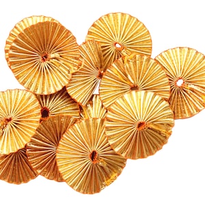 Puede incluir: Un grupo de ocho decoraciones circulares plisadas de color dorado con un pequeño agujero central naranja. Las decoraciones están dispuestas en un racimo.