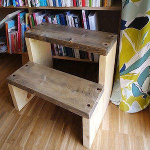 May include: A wooden step stool with a natural wood finish. The stool has two steps and is sitting in front of a bookshelf filled with books. The bookshelf is against a wall with a colourful floral patterned curtain.