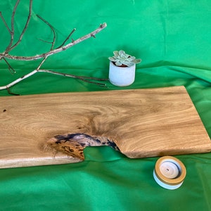 Könnte beinhalten: Ein Serviertablett aus Naturholz mit einer einzigartigen, unregelmäßigen Form und einem kleinen Loch in der Mitte. Das Tablett ist hellbraun und hat eine glatte, polierte Oberfläche. Es steht auf einer grünen Oberfläche mit einer kleinen Sukkulente im Topf und einem weißen Kerzenhalter.