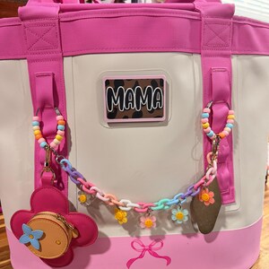 May include: A cream and pink tote bag featuring a rectangular patch with the text MAMA over a brown leopard print. The bag is decorated with a pastel-colored plastic chain, flower charms, a pink flower-shaped coin purse, and a pink bow graphic.