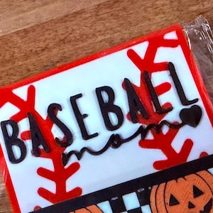 Może przedstawiać: Prostokątny znak z białym tłem i czerwonymi wzorami szwów baseballowych. Czarny napis brzmi BASEBALL mom z małą ikoną serca. Poniżej znajduje się czarno-biała szachownica umieszczona nad grafikami pomarańczowych dyń.