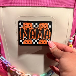 Könnte beinhalten: Ein rechteckiger Aufnäher mit dem Wort MAMA in schwarzen Buchstaben auf einem orange-schwarzen Kürbis- und Schachbrettmuster. Der Aufnäher ist auf einer weißen Fläche einer leuchtend pinken Tasche befestigt, die mit einer pastellfarbenen Kunststoffkette verziert ist.
