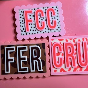 May include: Three rectangular name tags with scalloped pink borders. One tag features the letters FCC in coral on a white background with black dots. Another shows FER in white on a brown leopard print. The third displays CRUZ in coral on a red leopard print.