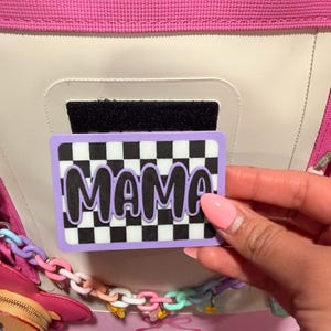 Puede incluir: Un parche rectangular con un fondo de cuadros blancos y negros y la palabra MAMA en letras moradas con un borde negro. El parche tiene un borde morado claro y está colocado sobre un bolso blanco y rosa con una correa de eslabones pastel.