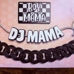 May include: A black and white keychain featuring the text DJ MAMA in white letters on a black background, attached to a thick black plastic chain and a silver metal ring. Above it is a rectangular black and white sign with the text BOY MAMA.