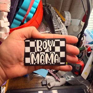 May include: A rectangular black and white checkered patch featuring the raised white text "BOY MAMA" in a rounded, bold font. The patch has a clean, high-contrast design with a matte finish, displaying the words clearly across the center.