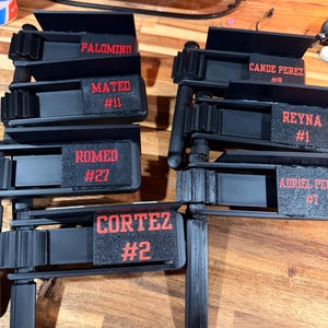 May include: A collection of black plastic noisemaker rattles, each featuring a textured rectangular panel with red printed names and numbers including Palomino, Mateo #11, Romeo #27, Cortez #2, Cande Perez #8, Reyna #1, and Adriel Perez #7.
