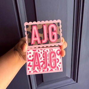 May include: Two rectangular plaques with scalloped edges featuring the letters AJG in raised pink text. The top plaque has a brown and black leopard print background, while the bottom plaque features a white and pink animal print pattern.