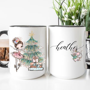 May include: Two white ceramic mugs with black handles. One mug features a ballerina with a Christmas tree and presents. The other mug has the name "heather" written in cursive and a mouse wearing a crown.