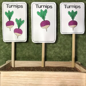 May include: Three wooden plant markers with white labels that say "Turnips" in black text. Each label has an embroidered image of a purple turnip with green leaves. The markers are in a wooden planter box with brown fabric lining.