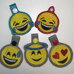 May include: Six embroidered keychains featuring different yellow smiley faces with various expressions. The keychains are made of felt and have a loop for attaching to keys.