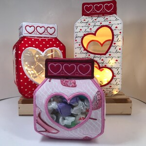 May include: Three decorative jar-shaped lights with heart cutouts and lids. The jar on the left is red with white polka dots. The middle jar is white with red hearts and the words "love, live, laugh". The jar in the front is pink with shoe graphics.