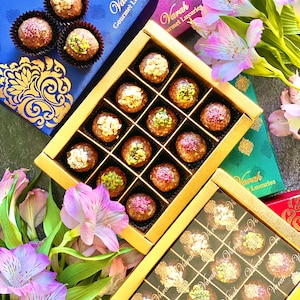 May include: Two gold boxes filled with gourmet chocolates. The boxes are decorated with the brand name "Varah Gourmet Luxuries". The boxes are surrounded by pink flowers.