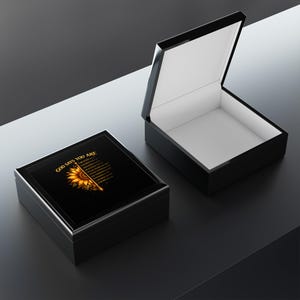 Puede incluir: Caja de madera negra con acabado brillante, la tapa con un diseño de girasol y el texto "GOD SAYS YOU ARE". La caja está abierta, revelando un interior blanco, y se encuentra sobre una superficie oscura.
