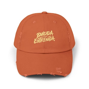 May include: An orange baseball cap with a distressed, worn look. The cap features the text "BANDIDA ENTRENADA" in a stylized, yellow font across the front. The hat has a curved brim and an adjustable strap.