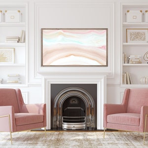 May include: Two pink velvet armchairs with gold metal frames sit in front of a fireplace with a white mantel and a black hearth. The fireplace has a black metal grate and a white surround. Above the fireplace is a framed abstract painting with pink, blue, and brown colors. The room has white walls and white built-in shelves with decorative objects and books.