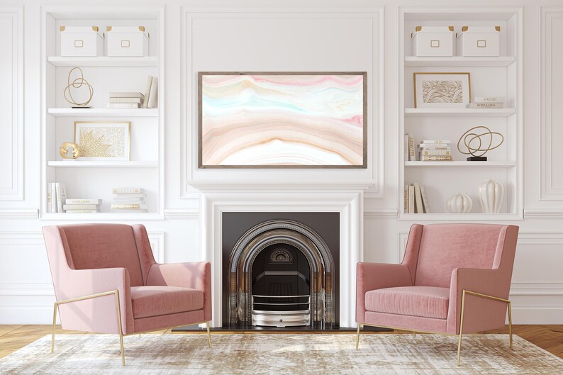 May include: Two pink velvet armchairs with gold metal frames sit in front of a fireplace with a white mantel and a black hearth. The fireplace has a black metal grate and a white surround. Above the fireplace is a framed abstract painting with pink, blue, and brown colors. The room has white walls and white built-in shelves with decorative objects and books.