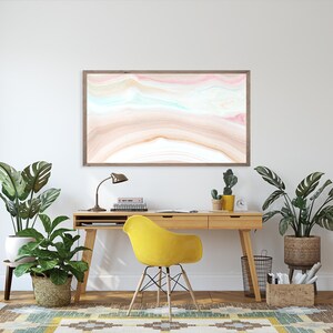 May include: A home office workspace with a wooden desk, yellow chair, and a framed abstract painting with pink, blue, and brown colors. There are two potted plants, a lamp, and a few books on the desk.