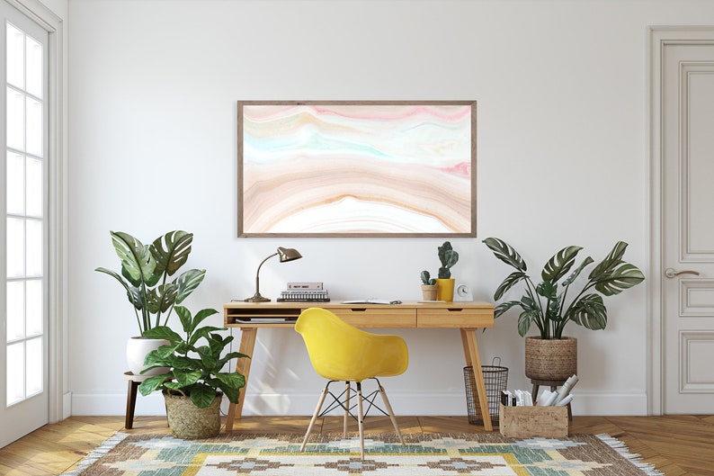May include: A home office workspace with a wooden desk, yellow chair, and a framed abstract painting with pink, blue, and brown colors. There are two potted plants, a lamp, and a few books on the desk.