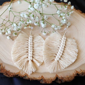 Puede incluir: Un par de pendientes de macramé color crema con diseño de pluma. Los pendientes están hechos de cordón de algodón anudado con una columna vertebral central tejida y bordes con flecos. Se exhiben sobre una superficie de madera con flores blancas.