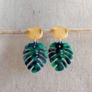 Puede incluir: Un par de pendientes con tachuelas ovaladas doradas y colgantes en forma de hoja verde y azul oscuro. Los pendientes están diseñados para parecerse a una hoja de monstera.