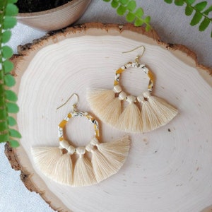 Peut inclure: Une paire de boucles d'oreilles à pompons crème avec des crochets dorés. Les cerceaux présentent un motif jaune et blanc. Les boucles d'oreilles sont présentées sur une surface en bois avec une plante en arrière-plan.