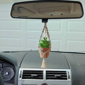 May include: A small, terracotta-colored pot with a green succulent plant hangs from a macrame plant hanger. The plant is suspended from a car's rearview mirror, adding a touch of nature to the vehicle's interior.