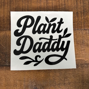 May include: A white sticker with the words "Plant Daddy" in a black, cursive font. The sticker has decorative leaf accents and is set against a wooden background.