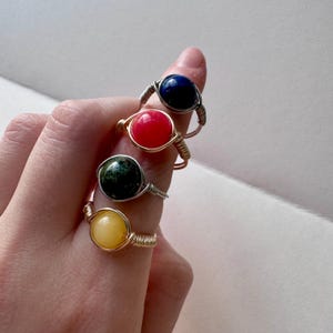 May include: Four wire-wrapped rings with colorful, round gemstones. The gemstones are blue, red, green, and yellow. The wire is silver and gold in color. The rings are displayed on a finger.