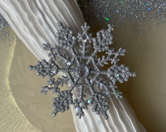 May include: A silver glitter snowflake napkin ring with a clear crystal in the center. The ring is on a white napkin.