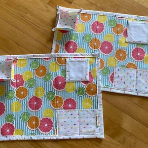 May include: Two rectangular placemats with a citrus fruit pattern. The placemats feature a blue and white striped background with colorful orange, pink, yellow, and green citrus slices. Each placemat has a small pocket and a white square patch.
