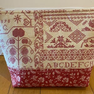 May include: Cream-colored fabric pouch with a red cross-stitch design. The pouch features a zipper closure, a wrist strap, and a red floral pattern at the base. Text on the pouch reads "what busy hands can do" and "ABCDEFGH".