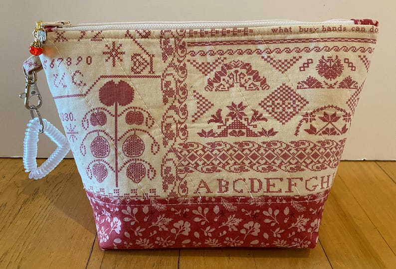 May include: Cream-colored fabric pouch with a red cross-stitch design. The pouch features a zipper closure, a wrist strap, and a red floral pattern at the base. Text on the pouch reads "what busy hands can do" and "ABCDEFGH".