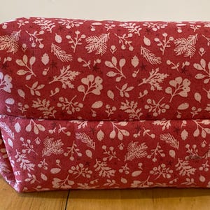 May include: A rectangular, red fabric pouch with a white floral pattern. The pouch has a zipper pull and is sitting on a wooden surface. The floral pattern includes leaves, berries, and other botanical elements.