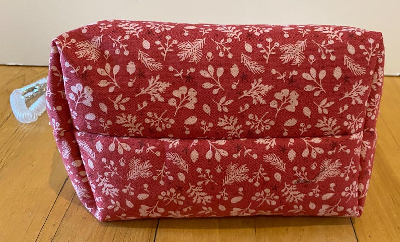 May include: A rectangular, red fabric pouch with a white floral pattern. The pouch has a zipper pull and is sitting on a wooden surface. The floral pattern includes leaves, berries, and other botanical elements.