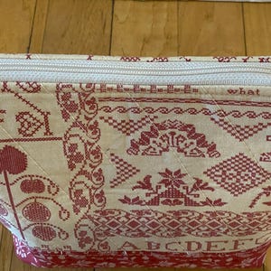 May include: A cream-colored, quilted cosmetic bag with a red cross-stitch design. The bag features a zipper closure and a decorative charm. The design includes floral and geometric patterns, along with the text "what busy hands can do."